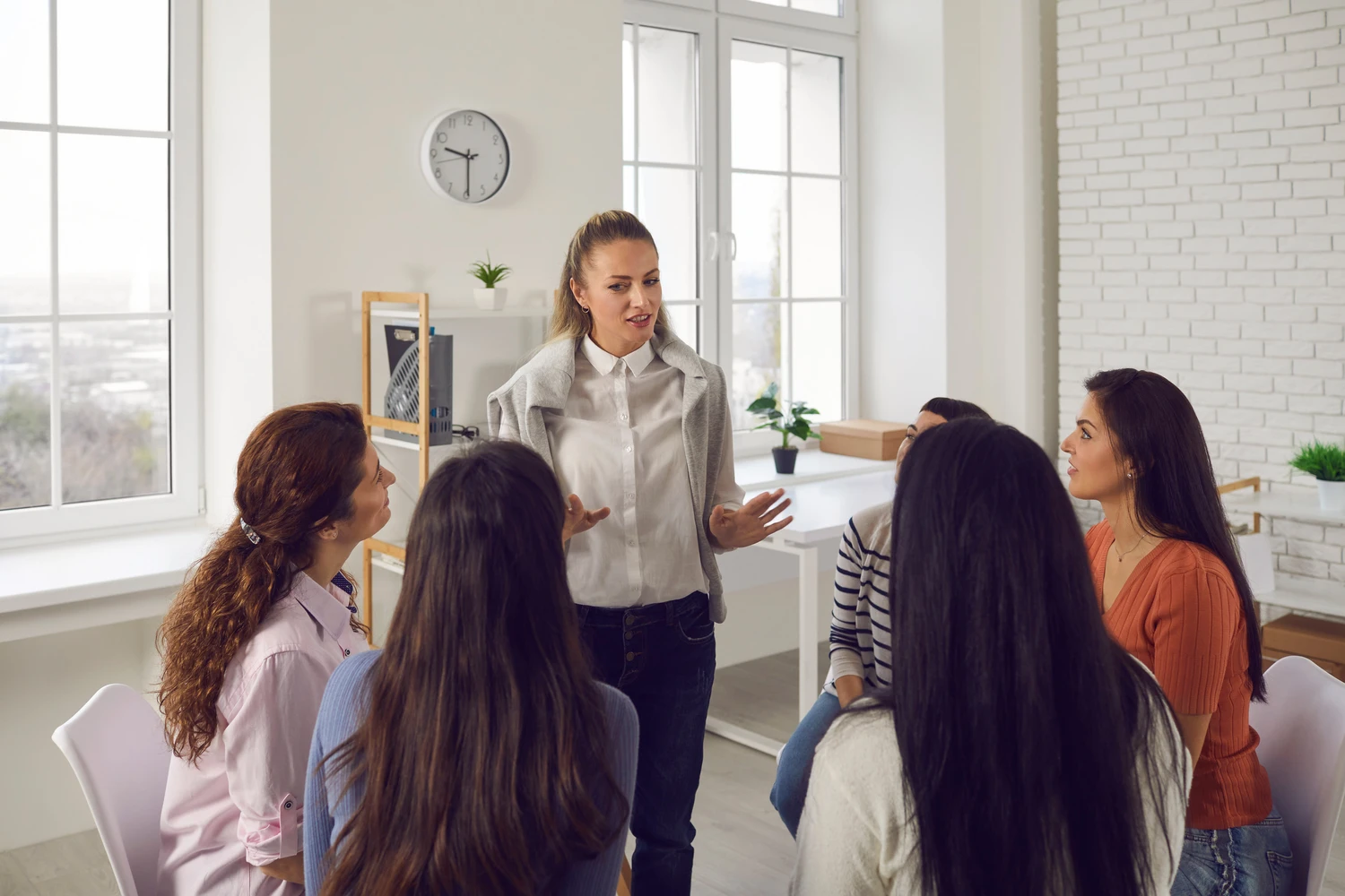 A woman stands and gestures while addressing a group of attentive listeners in a bright, modern room with large windows and plants